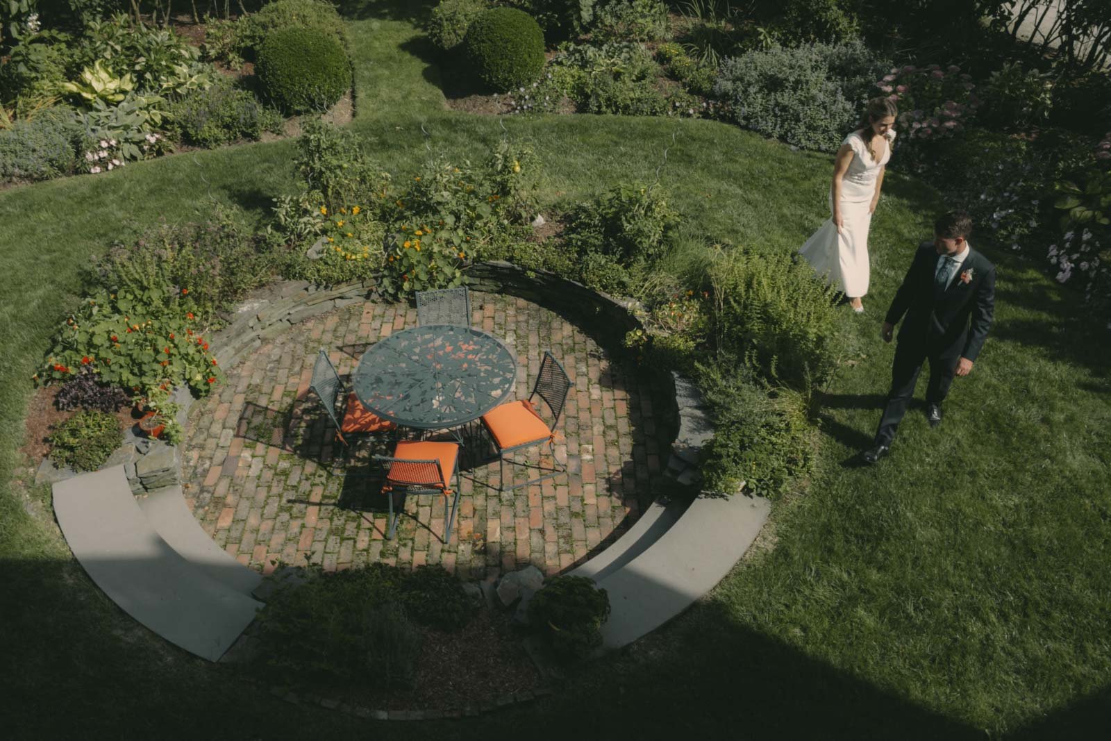 A man and woman in formal attire walking on a garden lawn near a table with chairs, surrounded by lush greenery and blooming flowers.