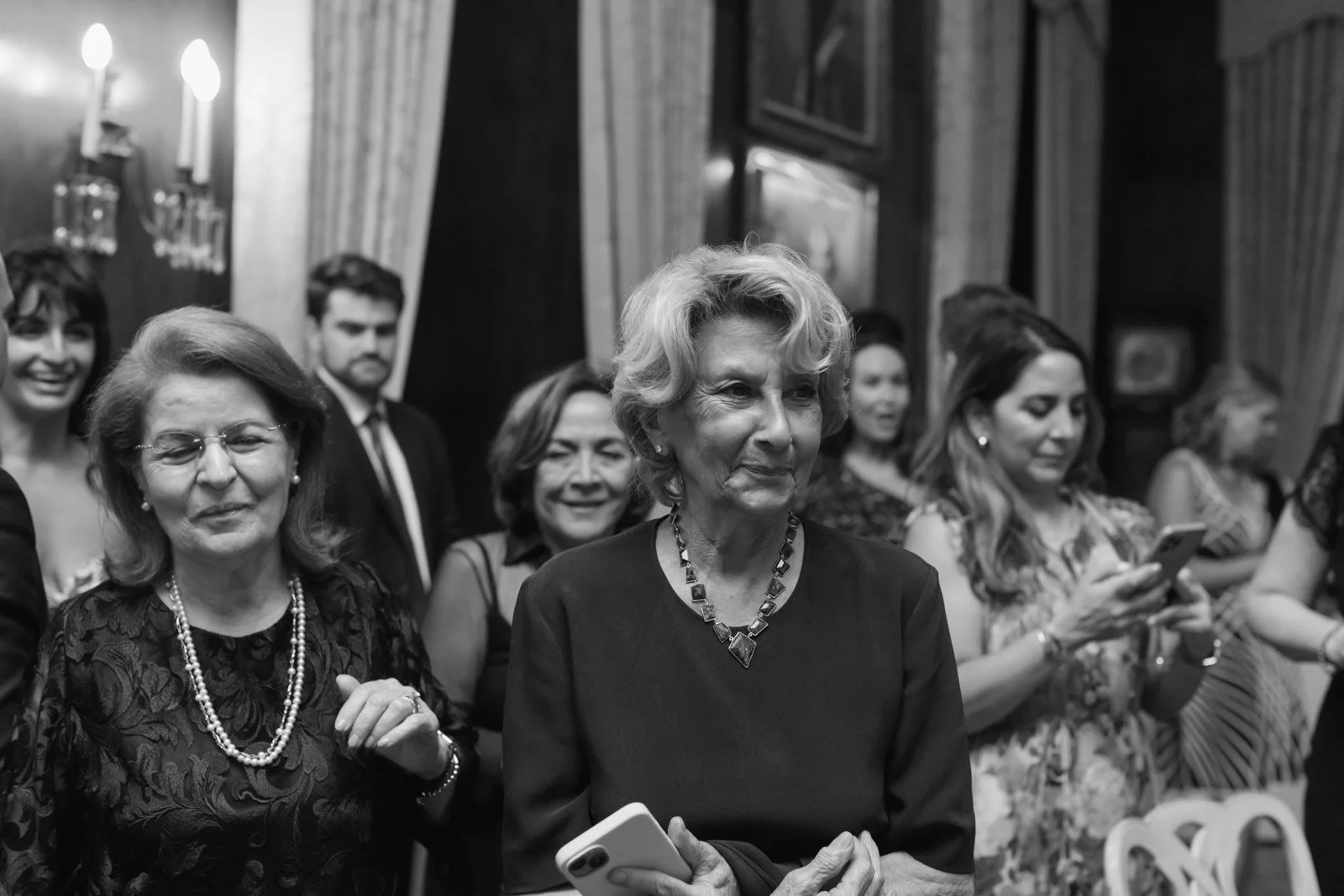 A group of women and a man attending an indoor event, with some women holding and looking at their phones, while others are smiling or watching something off-camera.