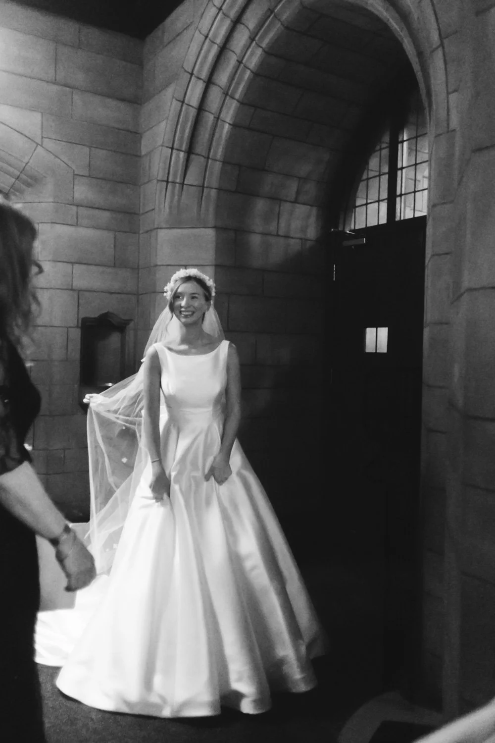 A bride in a white wedding dress and veil smiling in a church with stone walls and large arched window.
