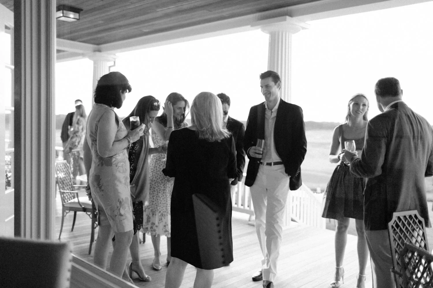 A group of people socializing on a covered porch at an outdoor gathering or party, with some holding drinks and engaged in conversation.