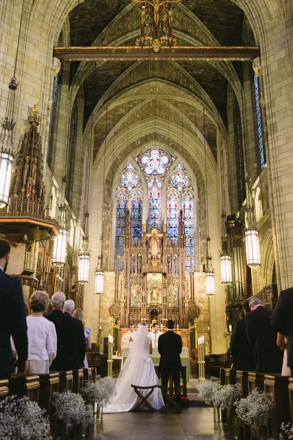 A wedding ceremony inside a grand Gothic-style church with stained glass windows, high vaulted ceilings, and ornate altar. Bride and groom are facing the officiant at the altar, surrounded by seated guests and floral arrangements.
