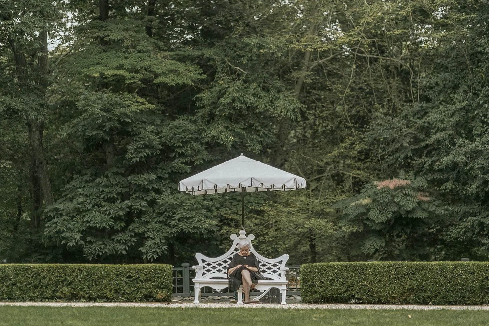 A woman sitting on a white bench under a white umbrella in a park with tall green trees and manicured bushes.