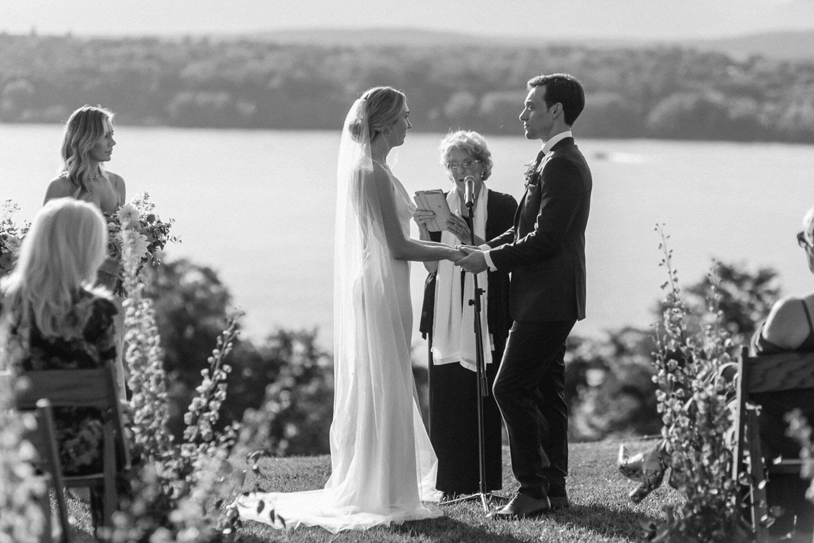 Black and white photo of a outdoor wedding ceremony with a bride and groom holding hands, exchanging vows, and an officiant reading from a book. Bridesmaid and guests are visible, with a scenic water view in the background.