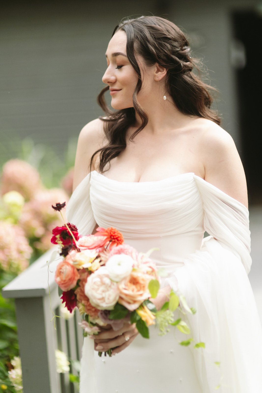 Brunette bride with wavy hair wearing off-the-shoulder white wedding dress holding a bouquet of pink and peach flowers, standing outdoors.