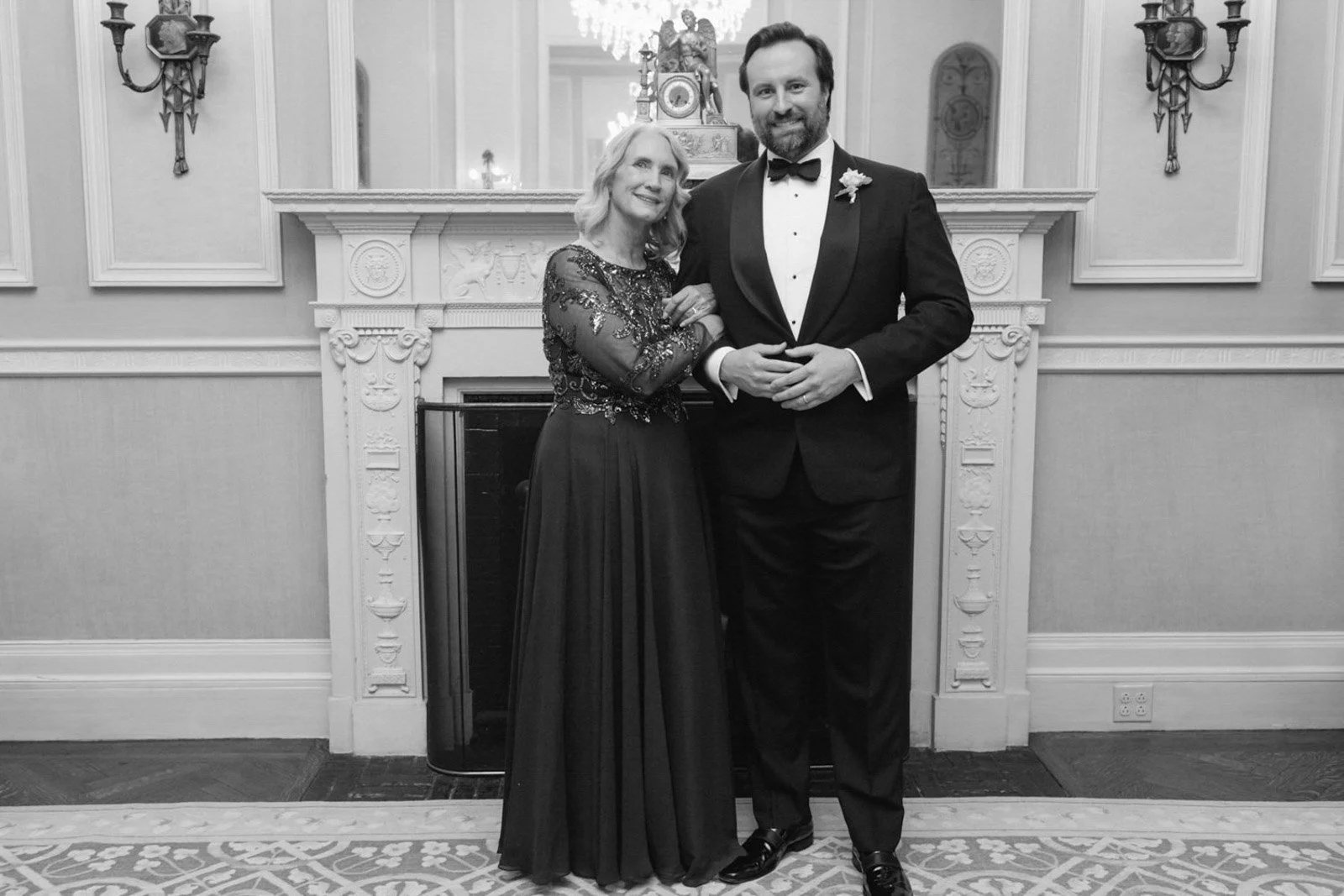 A woman and a man in formal attire posing in front of a fireplace with ornate fireplace mantle and mirror. The woman wears a long dress with embroidery, and the man wears a tuxedo with bow tie and boutonniere at The Colony Club New York.