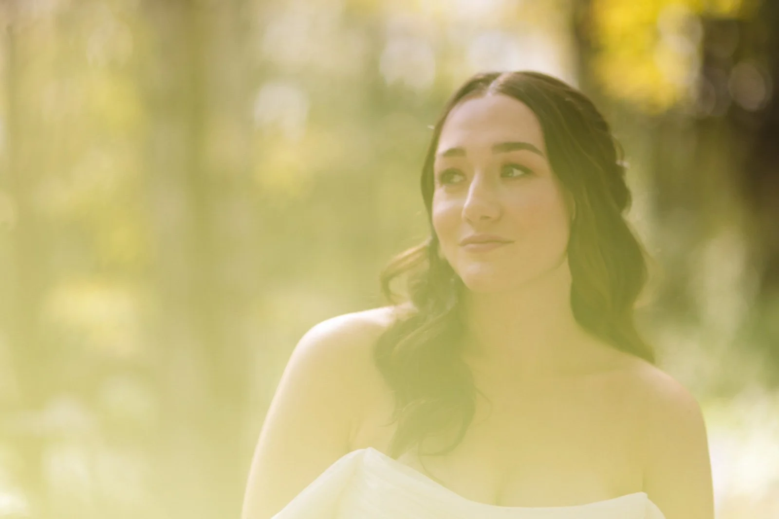 A woman with brown wavy hair, wearing a white off-shoulder top, standing outdoors with blurred yellow and green background, looking to her right.