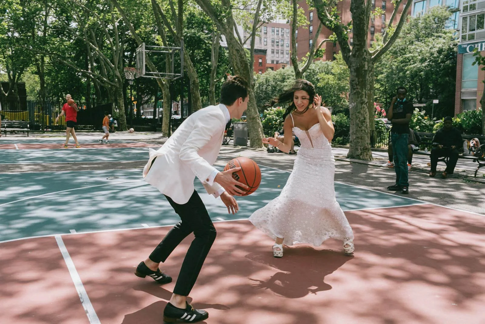 A man in a white blazer and black pants playing basketball with a woman in a white dress on an outdoor court in a park.