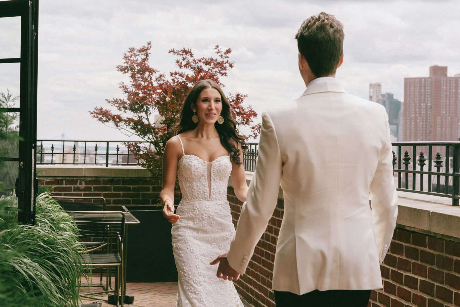 A woman in a white wedding dress holding hands with a man in a white suit on a balcony with city buildings in the background.