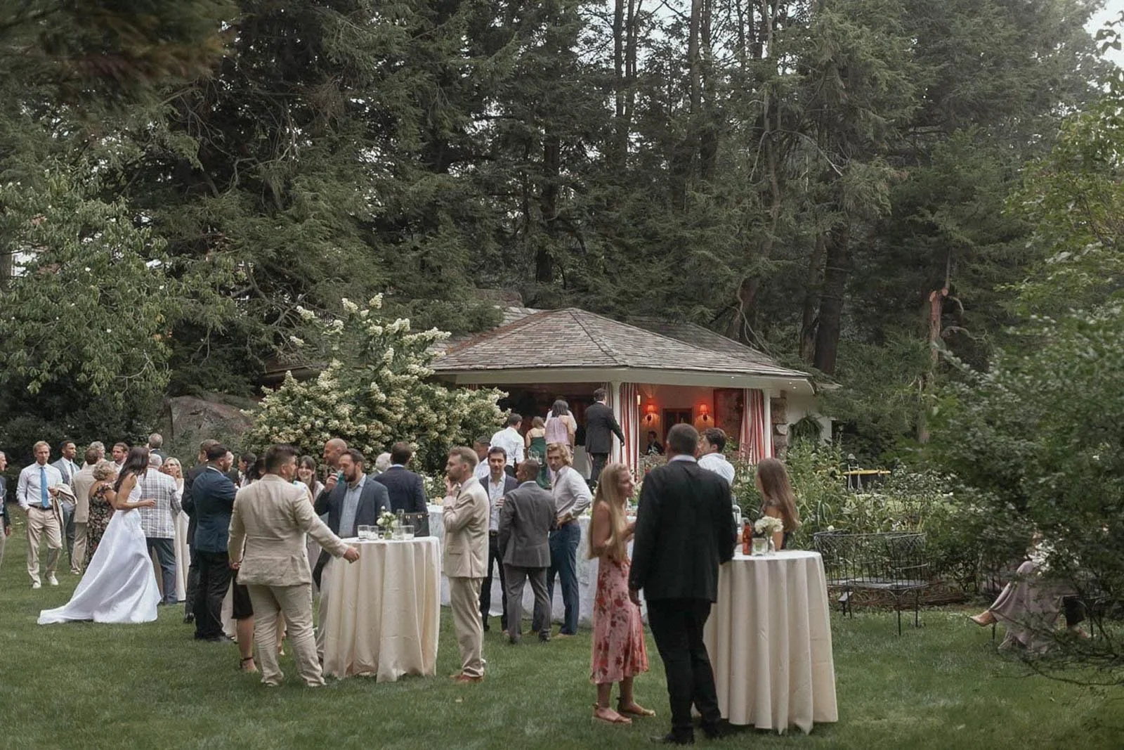 People gathered outdoors at a wedding reception or event in a garden with trees, a small building, tables, and floral decorations.