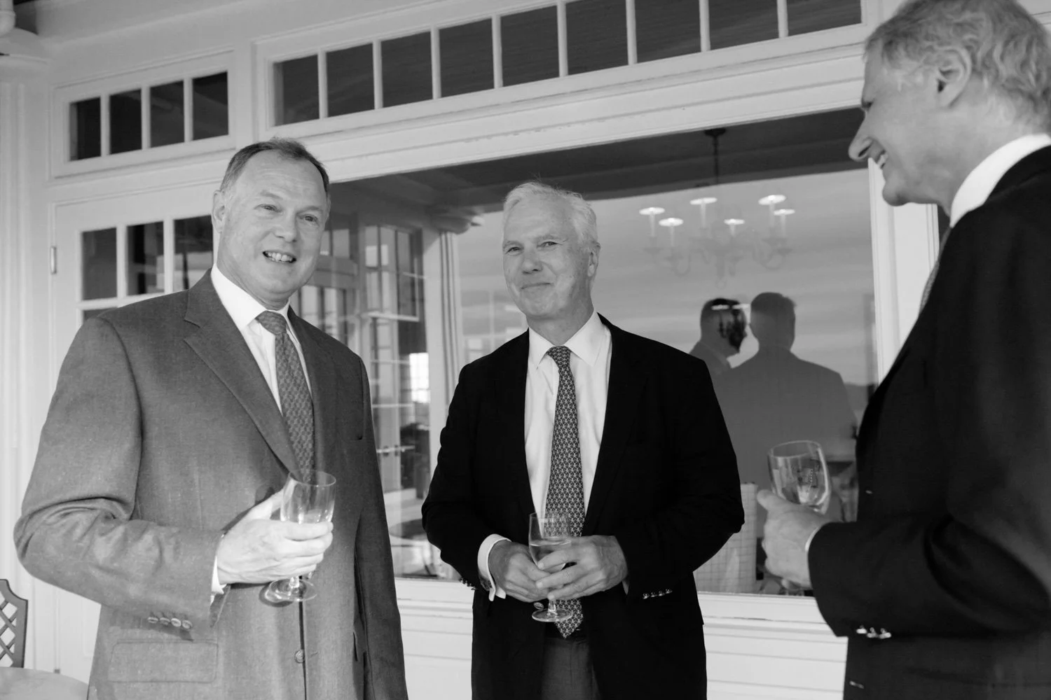 Three men in suits holding glasses and talking at a social gathering indoors, with large windows and chandeliers in the background.