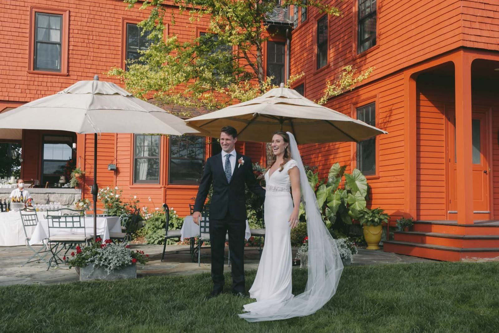 A newlywed couple standing together outdoors at a wedding reception, with the groom in a suit and the bride in a white wedding dress with a long veil. They are smiling, and there are large umbrellas, tables, and flowers in the background, with a red 
