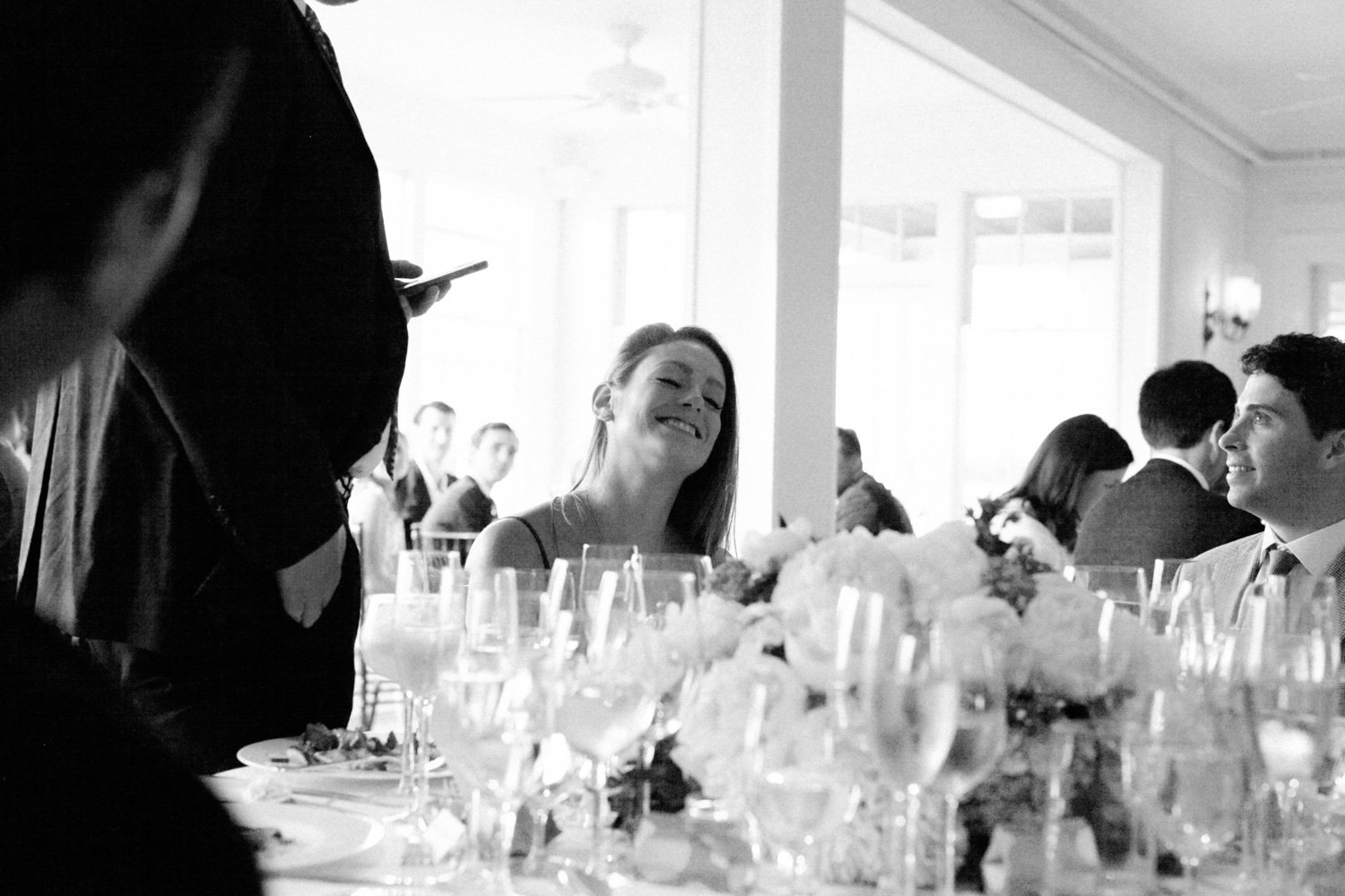 Smiling woman at a formal dinner in a brightly lit room with large windows, surrounded by glasses and floral centerpieces.