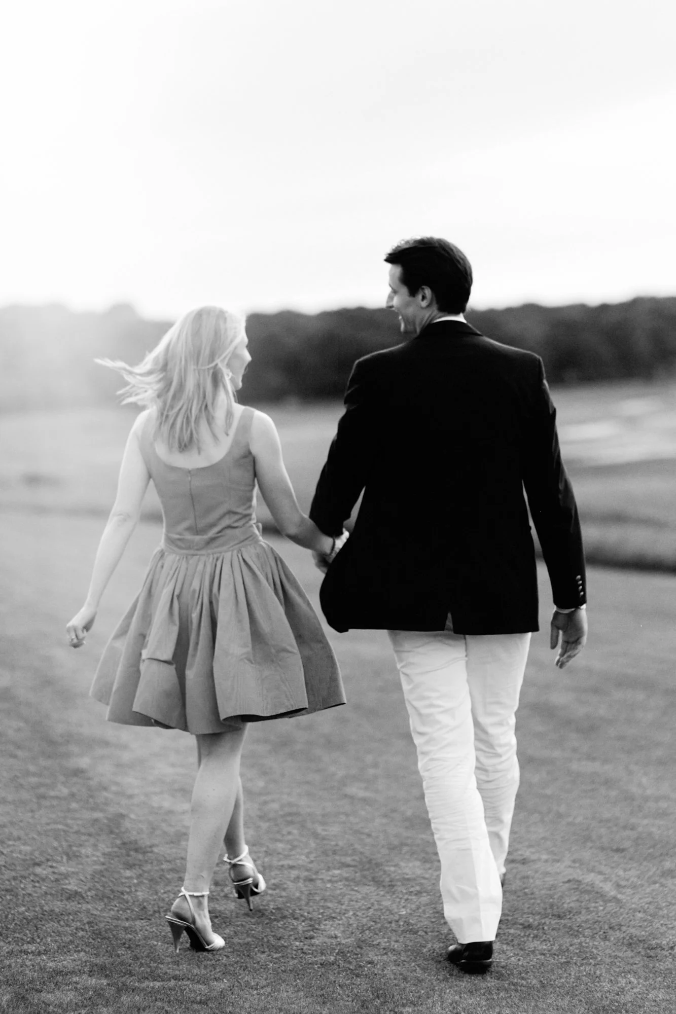 A black and white photo of a man and woman walking hand in hand outdoors, smiling and looking at each other, with a blurred landscape in the background.