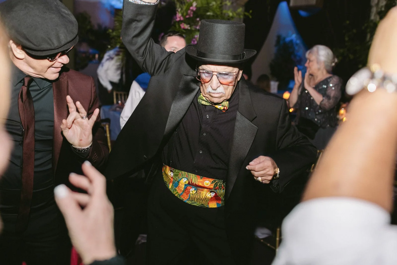 Older man wearing glasses, a top hat, a black suit, and a colorful cummerbund, dancing at a party with others in the background.