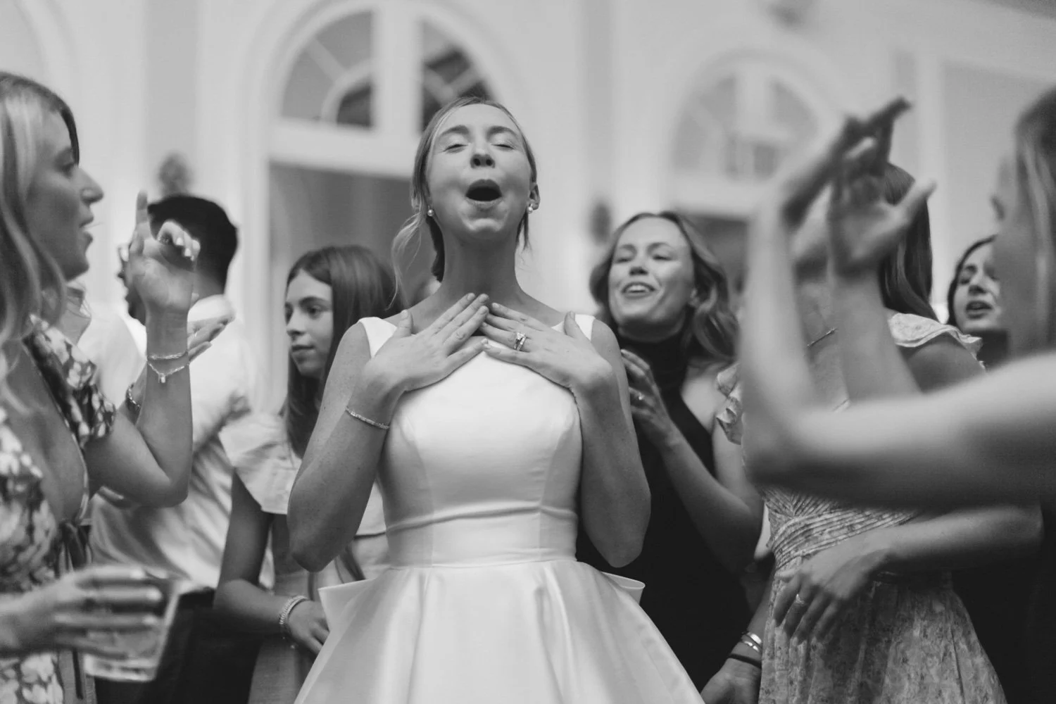 A woman in a white dress appears to be singing or speaking passionately with her eyes closed, surrounded by a group of women who are clapping, smiling, and engaging with her at an indoor social gathering.