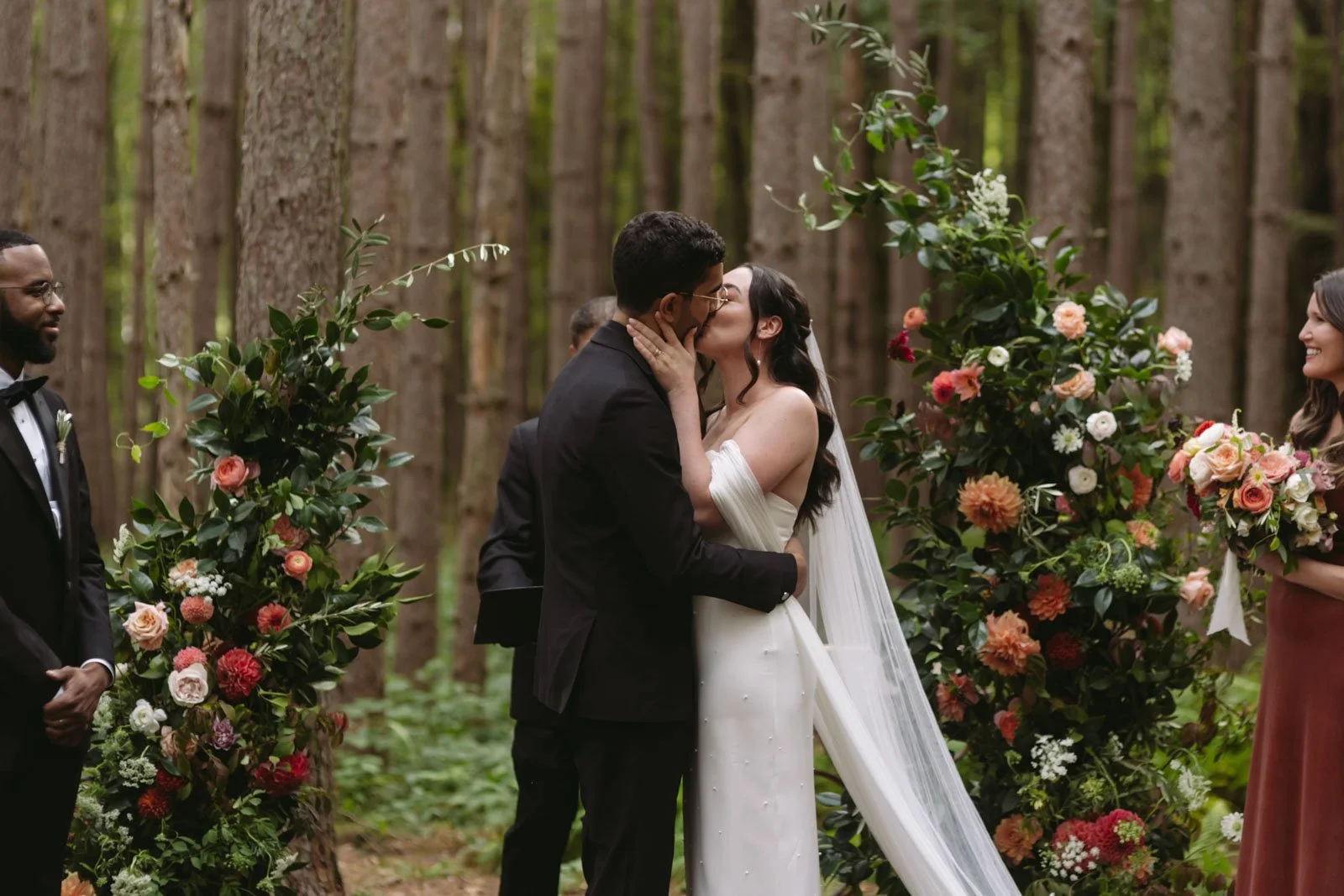A couple kissing during their outdoor wedding ceremony in a forest, surrounded by floral arrangements at Roxbury Barn and Estate.