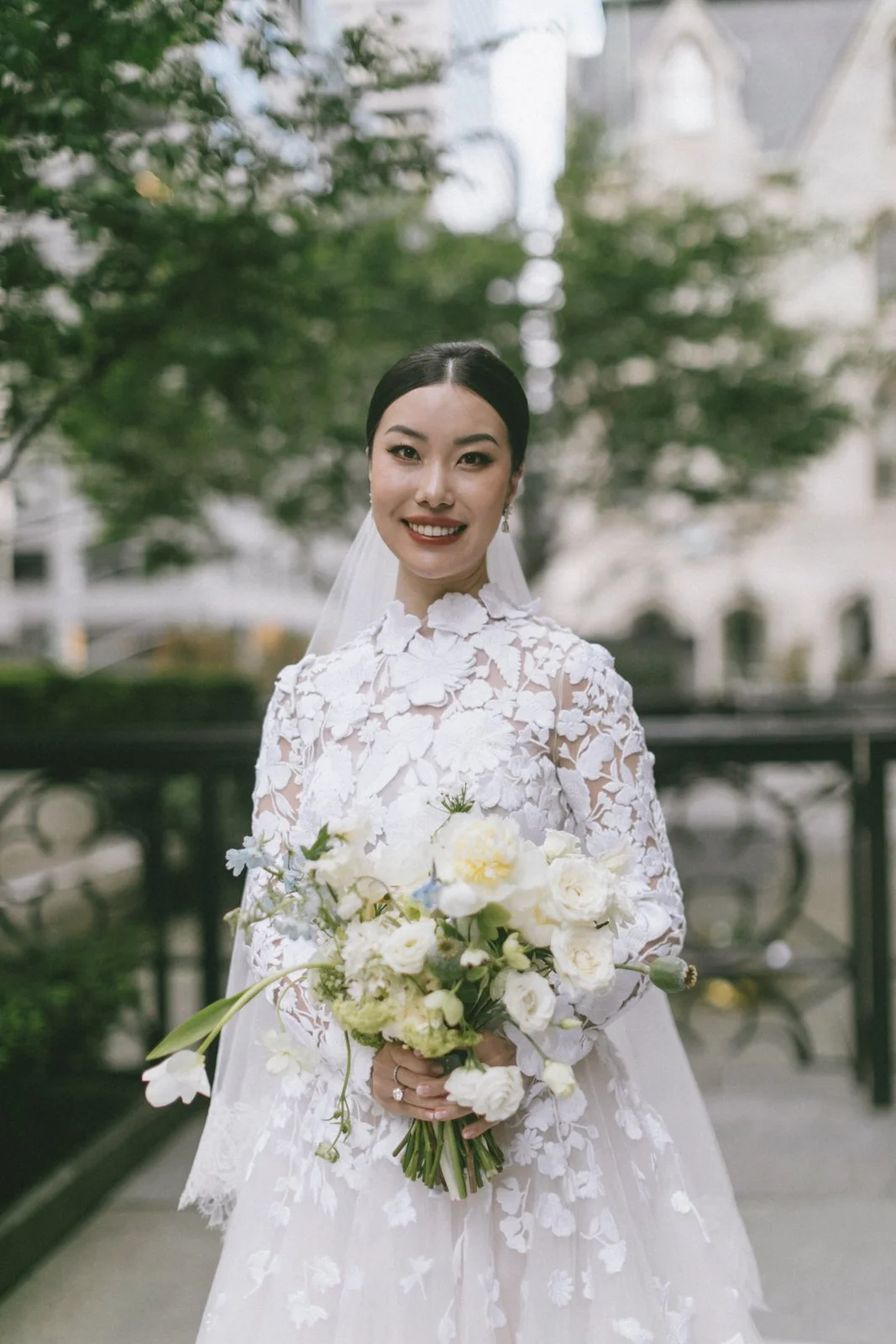 A smiling bride in a white wedding dress with floral lace details, holding a bouquet of white flowers, standing outdoors with trees and buildings in the background.