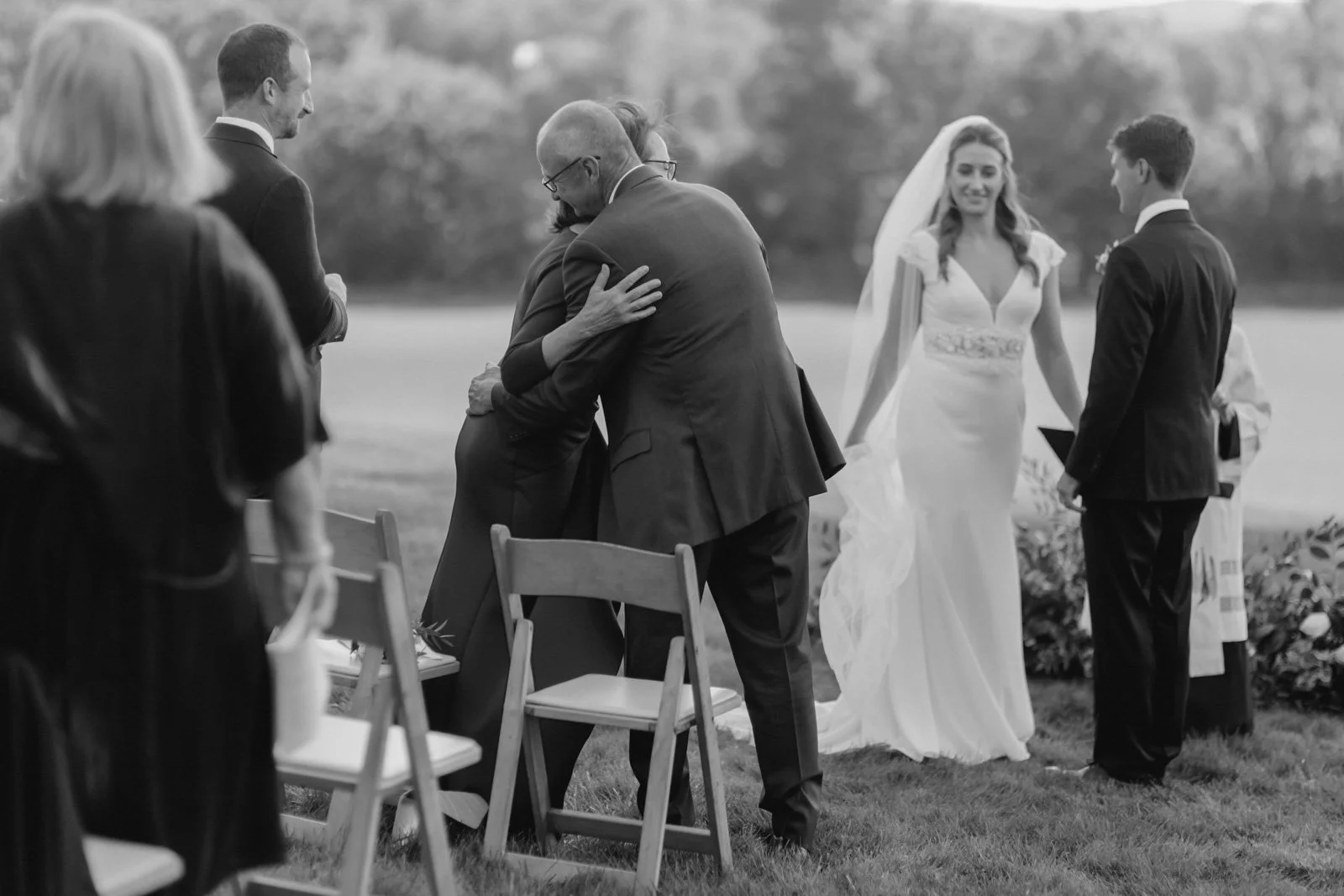 Black and white photo of a wedding ceremony outdoors, with a bride in a white gown and veil, a groom in a dark suit, and several guests.