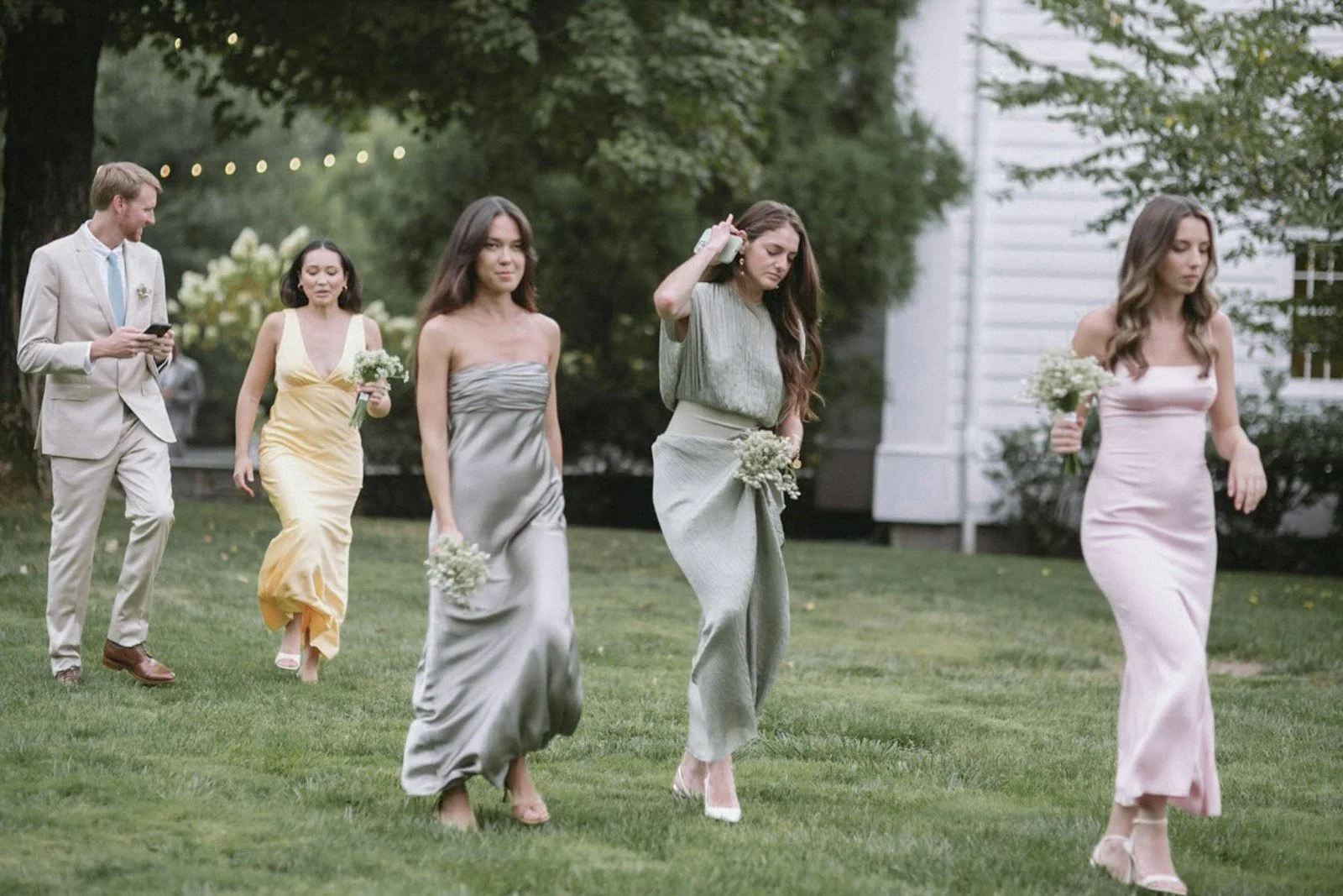 A group of six well-dressed people walking on a grass lawn outdoors, with three women in pastel-colored dresses carrying bouquets of flowers, and a man in a light suit looking at his phone, during a wedding or outdoor formal event.