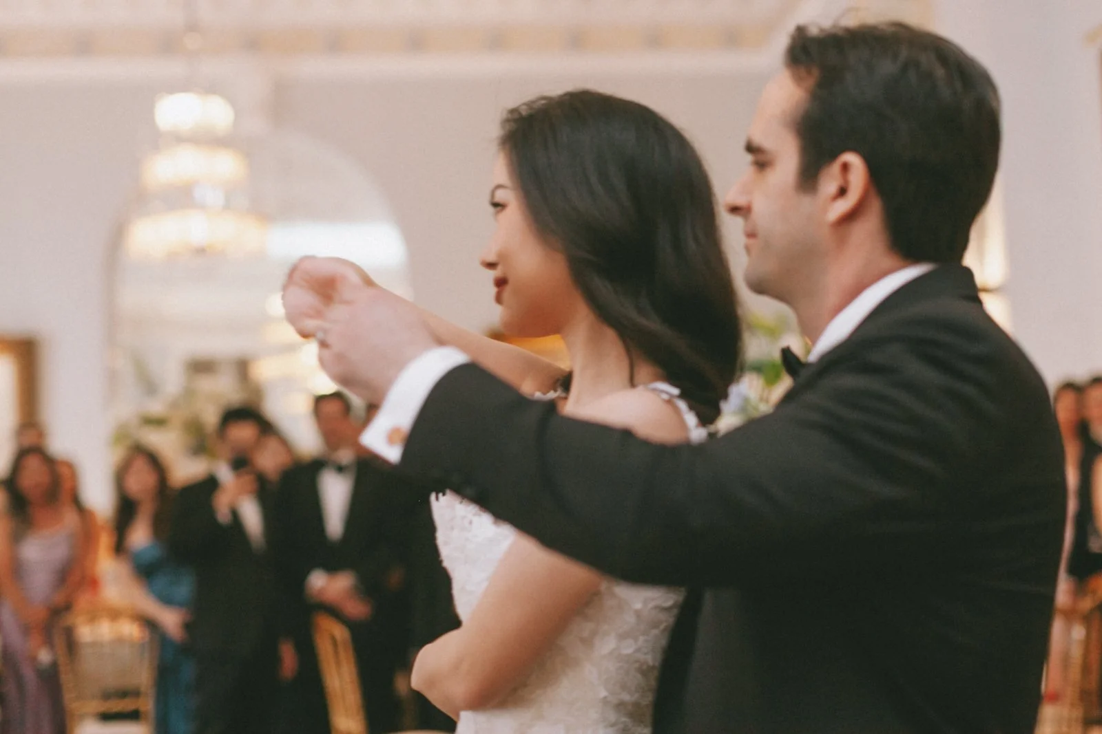 A bride and groom dancing at their wedding reception. The bride has dark hair and is wearing a white lace dress, while the groom has dark hair and is wearing a black tuxedo. Guests are visible in the background, watching the couple dance.