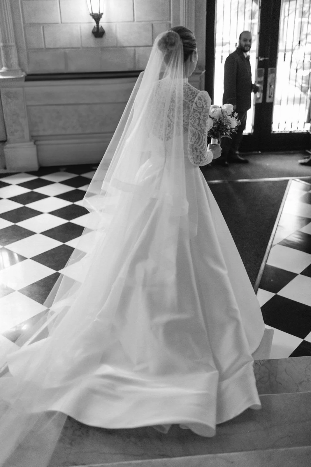 A bride in a wedding dress and veil holding a bouquet, standing inside a building near the entrance, with a man in a suit in the background.