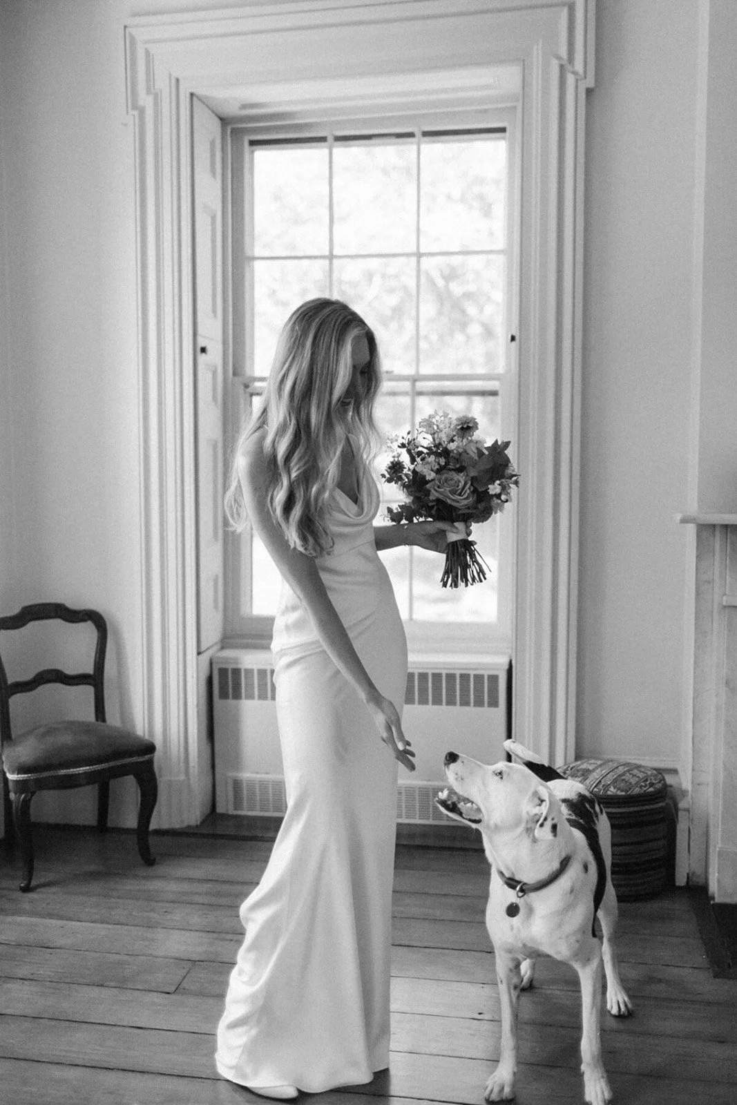 A black-and-white photo of a woman in a long dress holding a bouquet of flowers, interacting with a dog indoors near a large window.