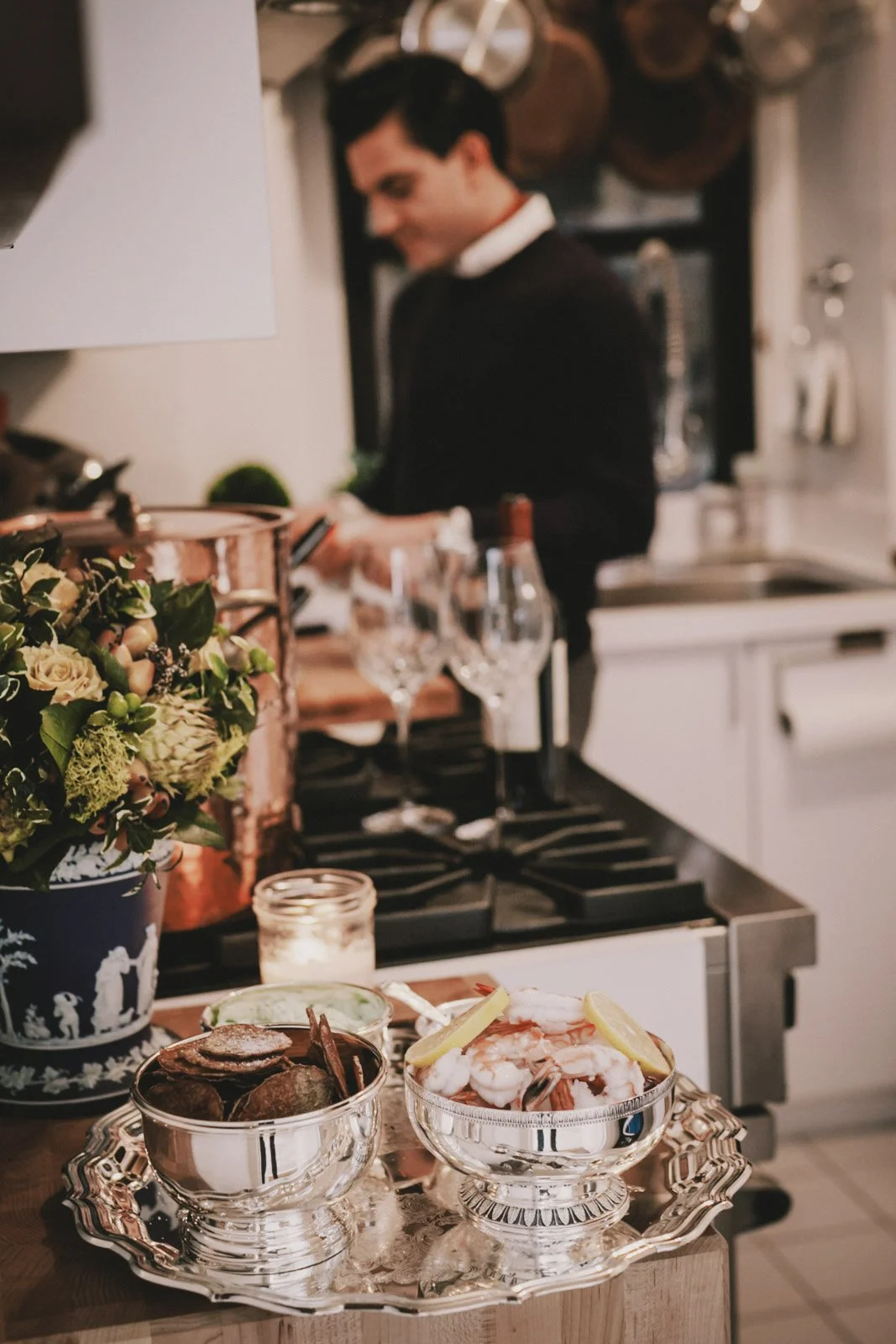 A chef is preparing food in a kitchen with a table in the foreground holding bowls of seafood garnished with lemon wedges, a candle, and a floral centerpiece.