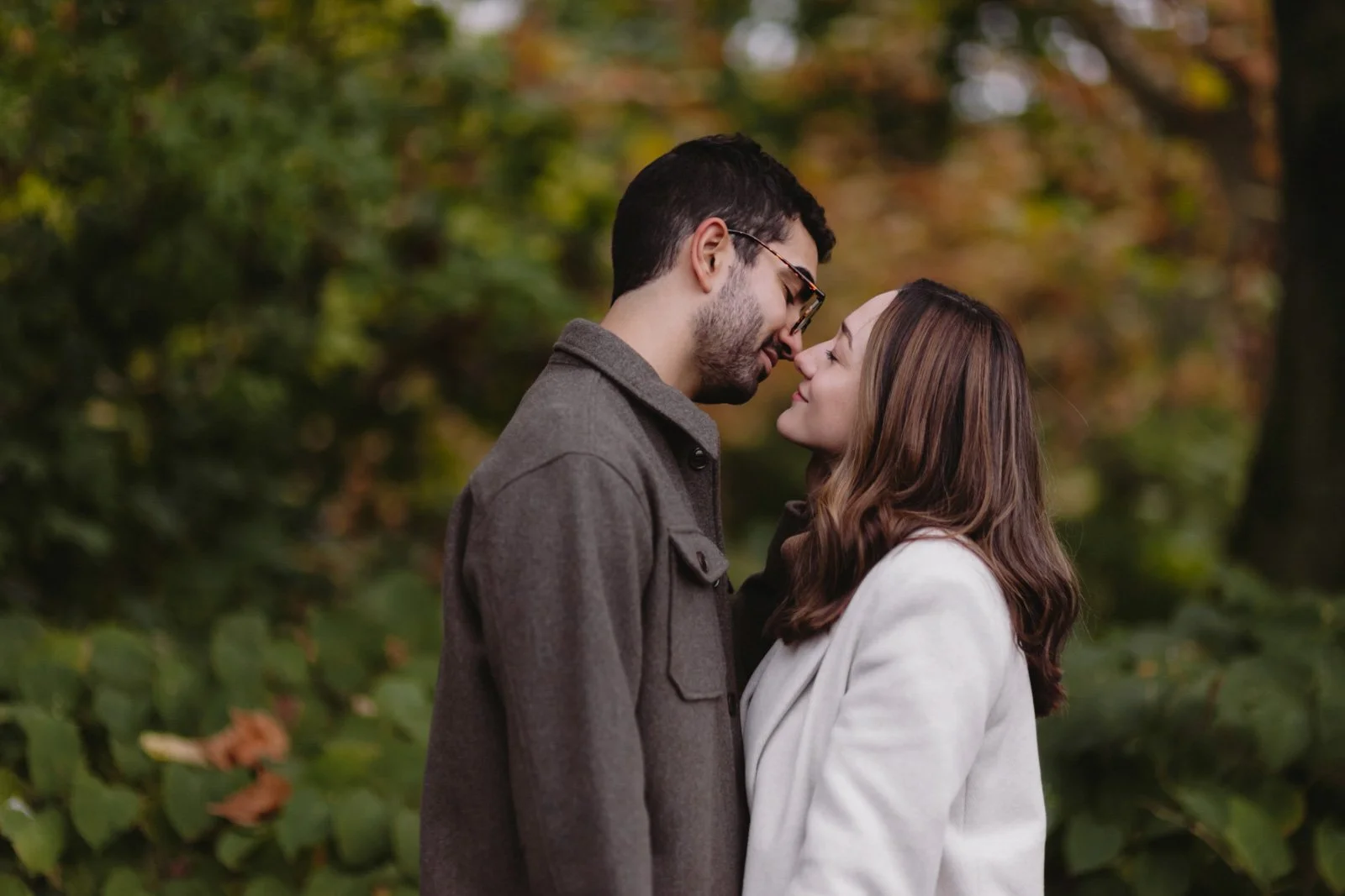 A couple standing close together outdoors, about to kiss, with blurred autumn foliage in the background.