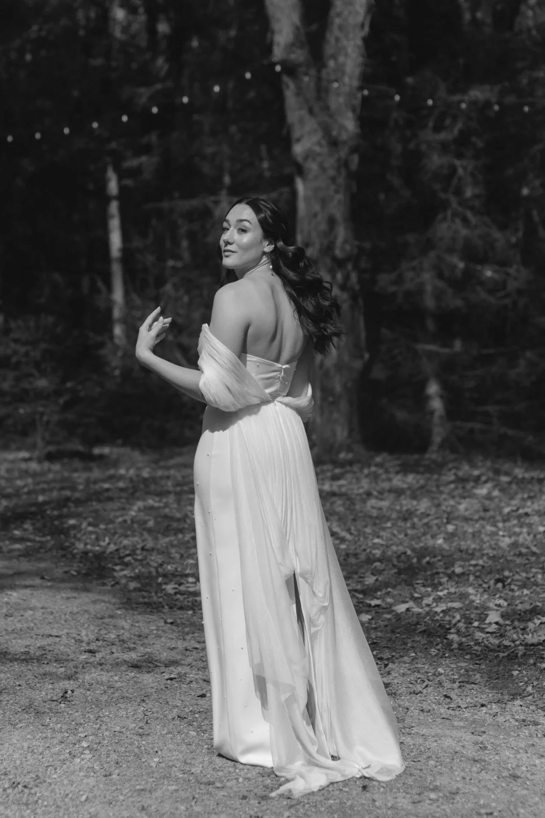 A woman in a long, elegant dress standing outdoors at night, with trees in the background, looking over her shoulder.