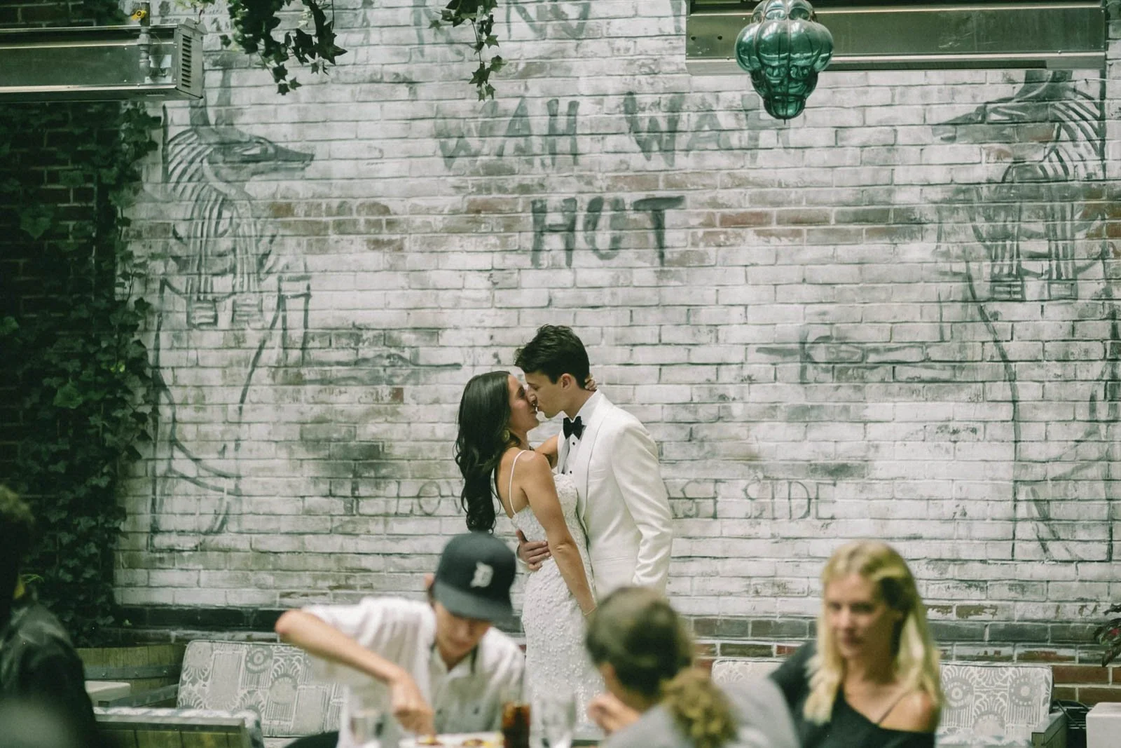 A couple in wedding attire sharing a kiss in front of a brick wall with "Wah Wah Hut" painted on it, surrounded by seated guests at a restaurant or bar.