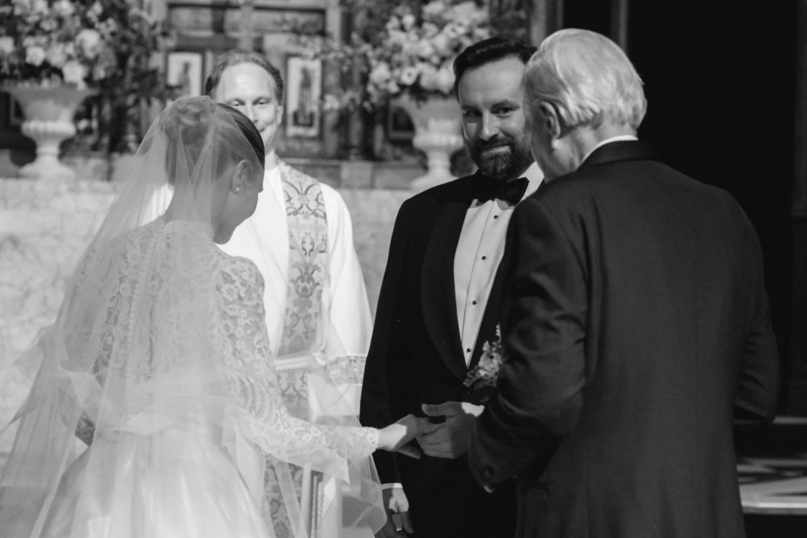 A wedding ceremony with a bride and groom holding hands in front of an officiant and an older man, in a church decorated with flowers and framed pictures.