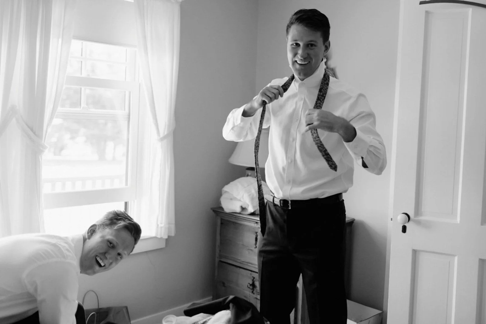 Two men preparing for a formal event in a bedroom. One man is sitting on the floor, smiling, and the other is standing, holding a necktie around his neck. The standing man is dressed in a white shirt and dark pants.