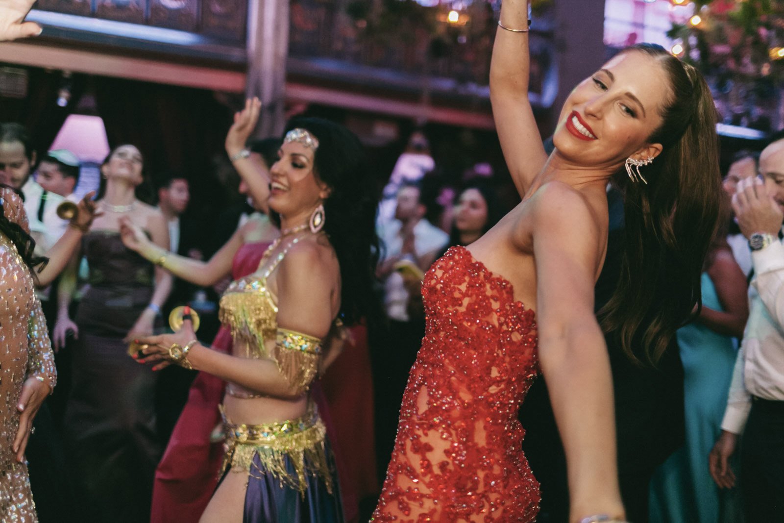 Group of women dancing at a party, dressed in glamorous outfits, with one woman in the foreground wearing a red embellished dress and smiling.