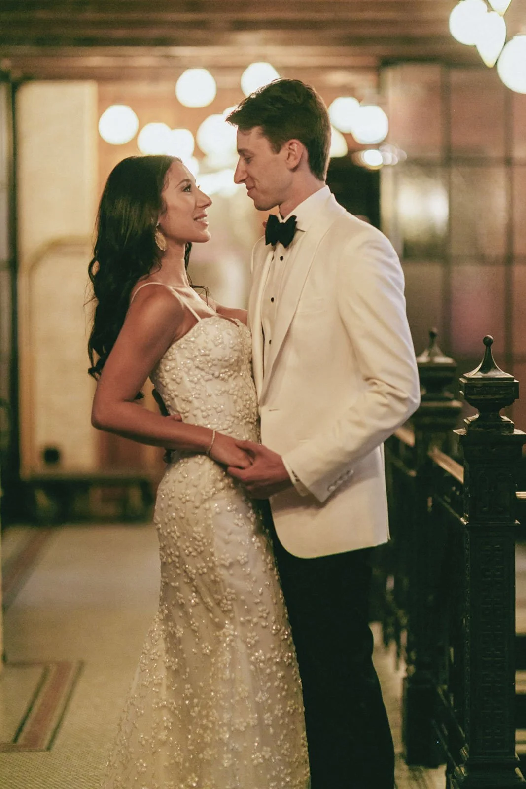 A man and woman in wedding attire, standing close and looking into each other's eyes in a warmly lit room.