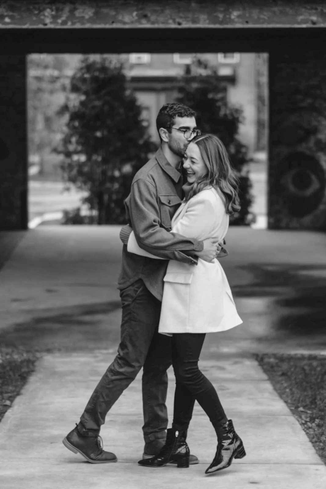 A black-and-white photo of a couple hugging and smiling on a sidewalk. The man is wearing glasses, a button-up shirt, and pants, while the woman is dressed in a coat and boots. They are standing under an archway with trees and a building in the backg
