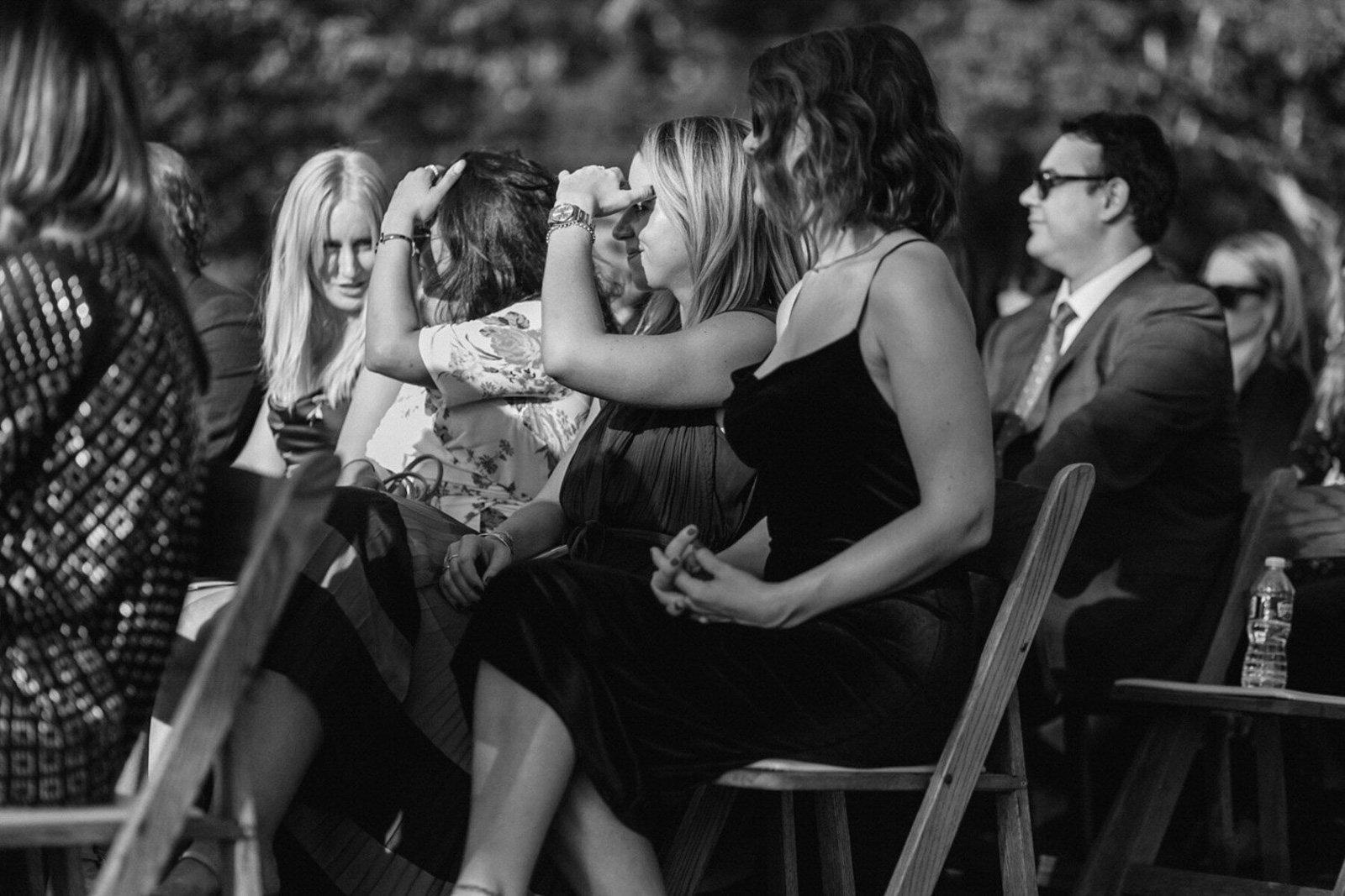 Black and white photo of people sitting on chairs at an outdoor event, some talking and others listening.