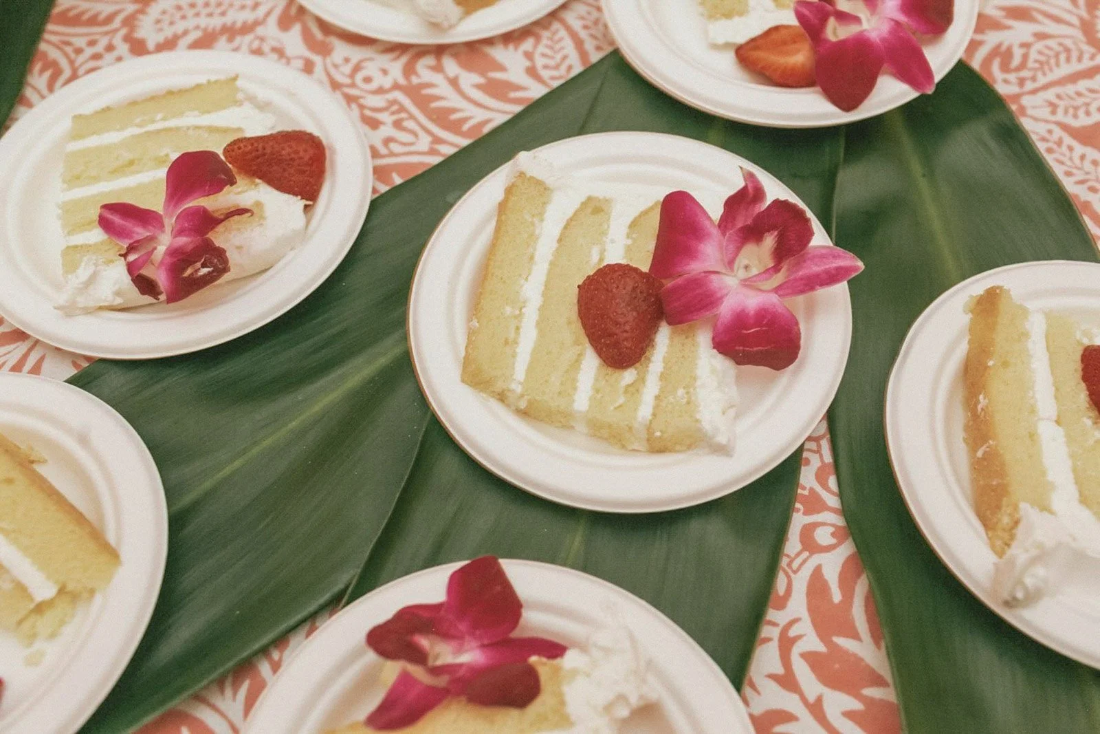 Several pieces of vanilla sponge cake with white frosting on small white plates, garnished with strawberries and pink orchid flowers, arranged on large green leaves.