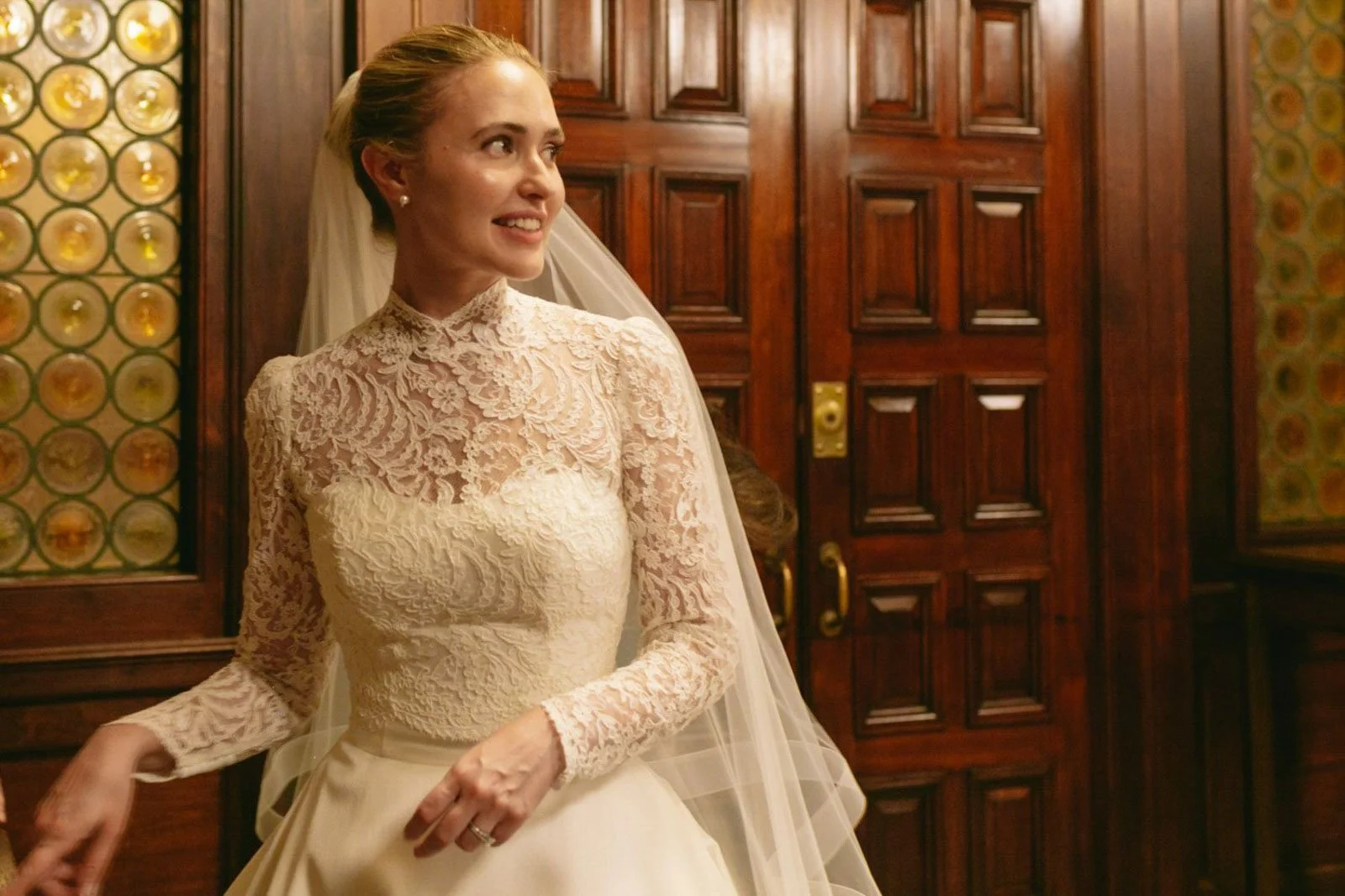 A smiling woman in a vintage wedding dress with lace long sleeves and a high neckline, standing inside a room with dark wood paneling and decorative glass panels.