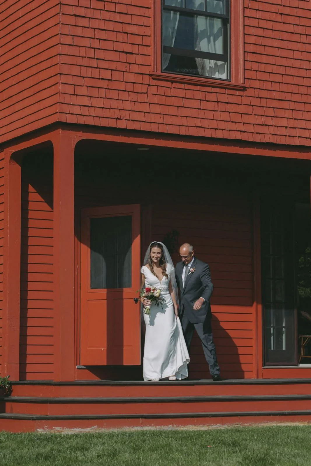 A bride in a white wedding dress holding a bouquet of flowers and smiling, walking out of a red wooden house with a man in a dark suit. They are standing on a porch with a grassy lawn in front.