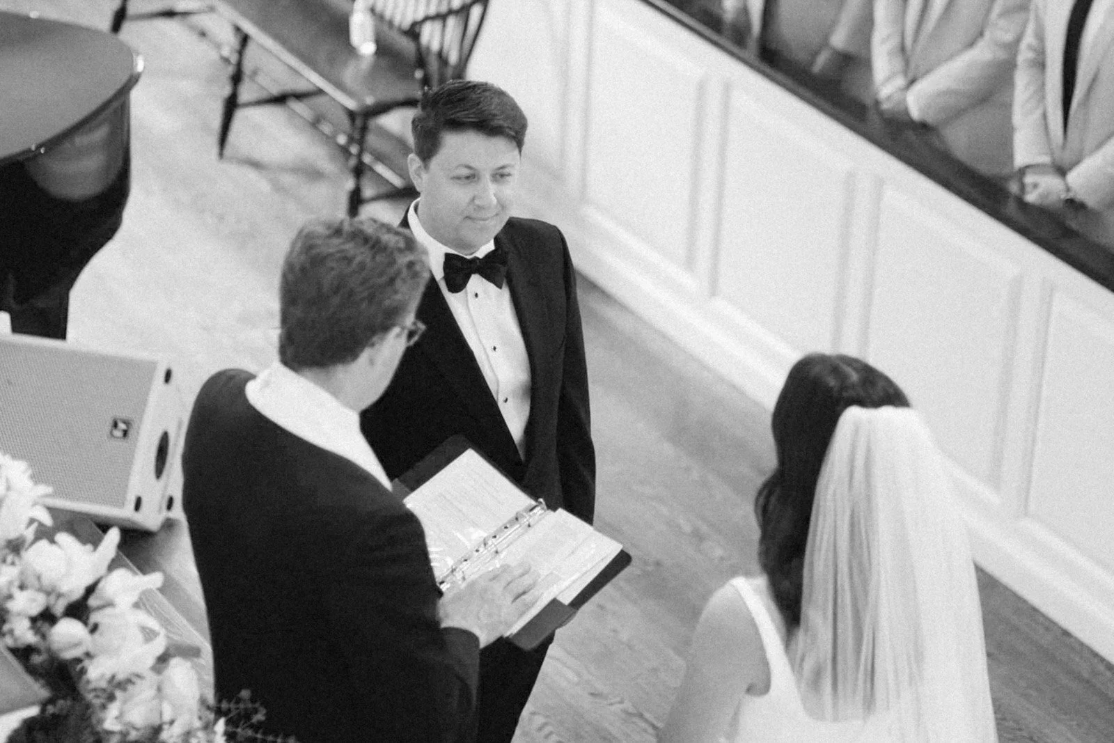 A black and white photo of a wedding ceremony with a groom in a tuxedo, a woman in a wedding dress, and a man in a tuxedo reading from a book.