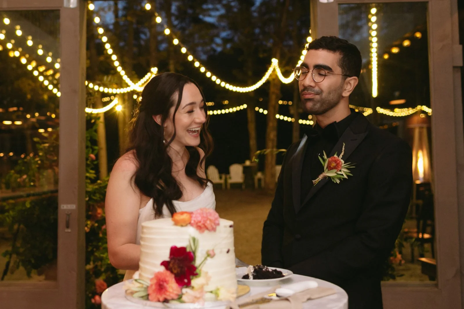 A newlywed couple at their wedding reception, standing at a cake table with a white wedding cake decorated with pink and orange flowers, indoors with string lights hanging in the background during the evening.