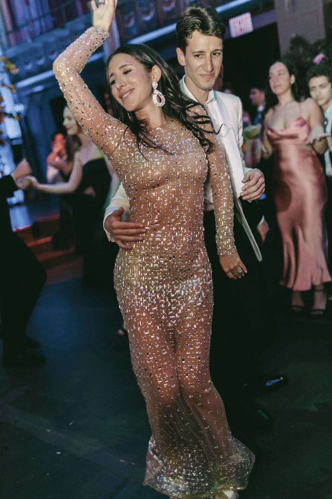 A woman in a long, nude-colored dress covered in shiny sequins dancing with a man in a tuxedo at a party or celebration.