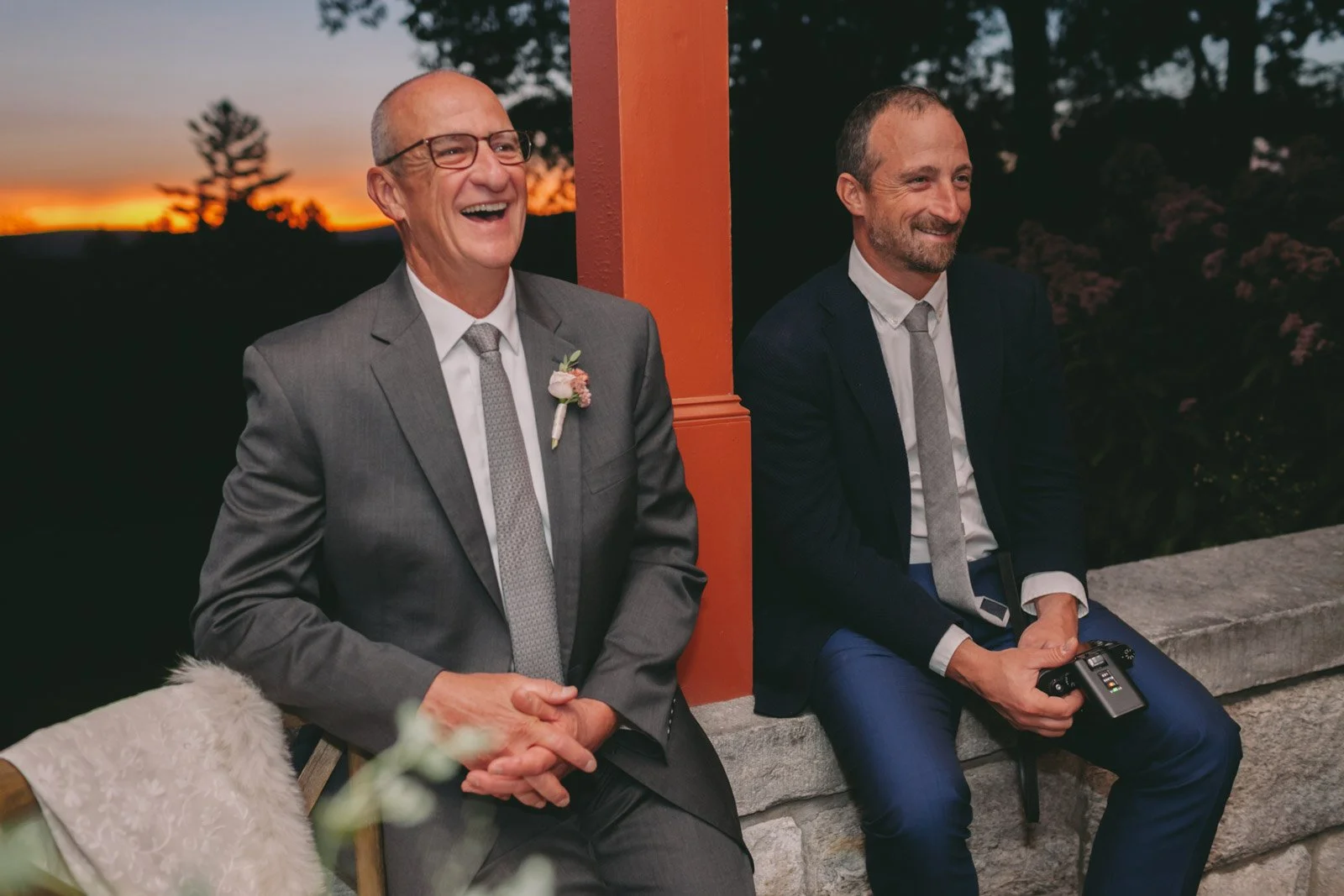 Two men in suits sitting outdoors during sunset, smiling and laughing, one holding a camera.