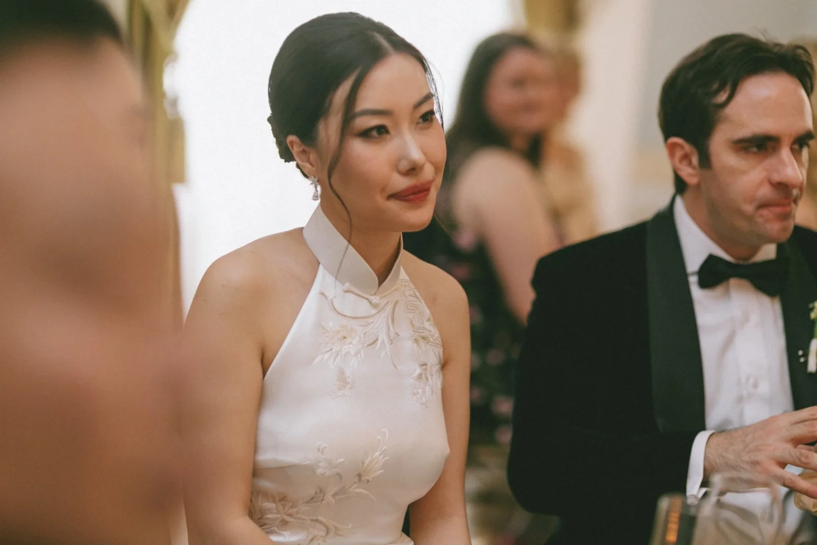 A woman in a white dress with floral embroidery and a high collar, sitting at a formal event with a man in a tuxedo. The woman has dark hair styled in an elegant updo with a loose curl framing her face. The man has dark hair, a bow tie, and a boutonn