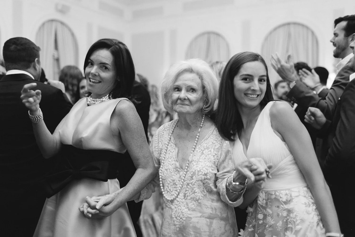 Three women are dancing at a formal event, holding hands and smiling. The woman in the middle appears elderly, while the other two are younger. They are dressed in elegant dresses, and the background shows more people dancing and socializing.