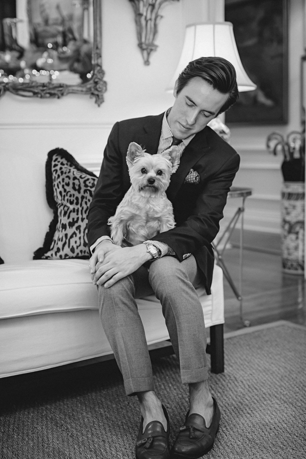 A man in a suit sitting on a sofa holding a small dog, in a cozy living room with a mirror, lamp, and decorative pillows.