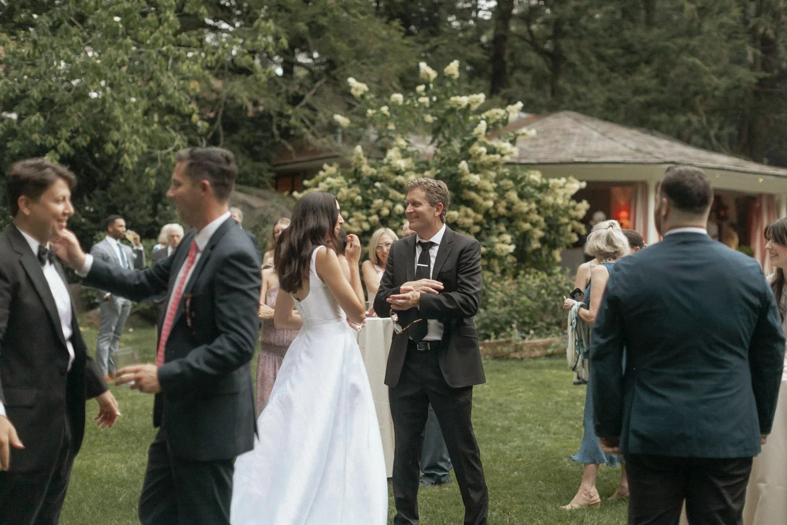 People socializing at an outdoor wedding reception, with women in dresses and men in suits, under trees and near a small building with a sunny ambiance.
