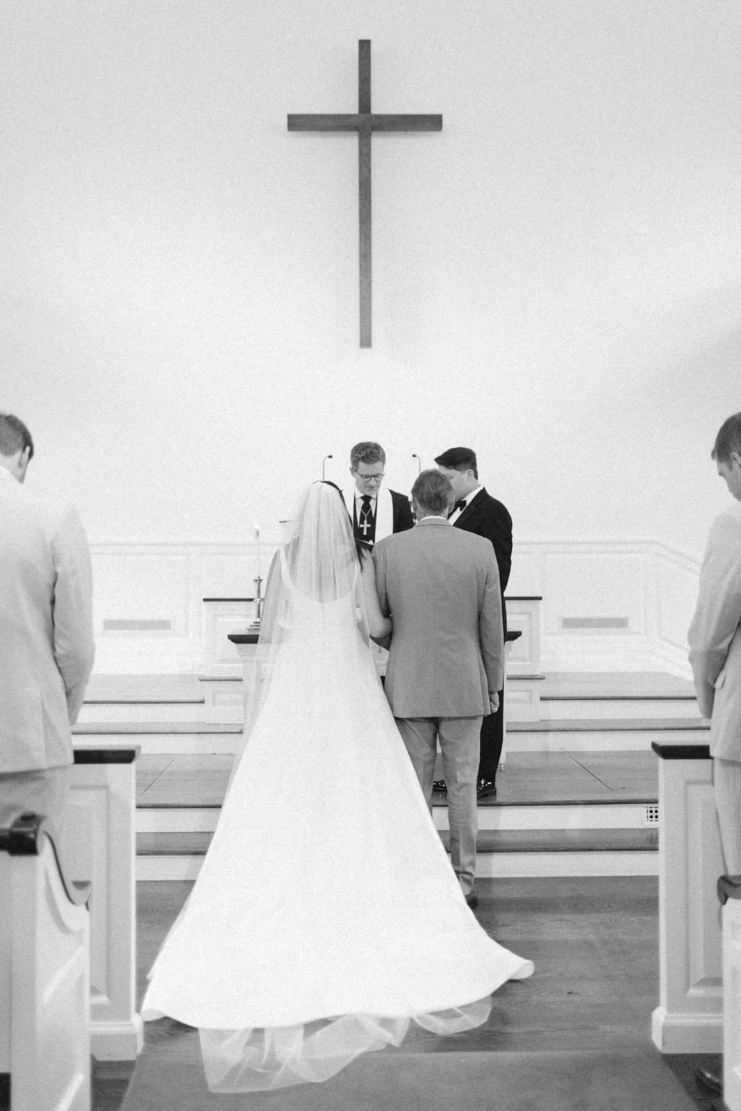 A black and white photo of a wedding ceremony inside a church, with a couple standing at the altar. The bride is in a long white gown with a veil, and the groom is in a light suit. Two officiants are assisting, while other people are watching. A larg