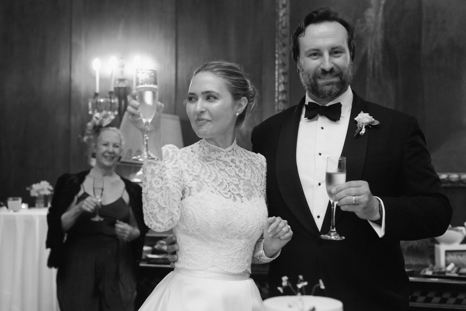 A black and white photo of a wedding reception with a bride and groom holding champagne glasses, smiling. The bride is in a lace dress, and the groom is in a tuxedo. A woman in the background is smiling and holding a glass.