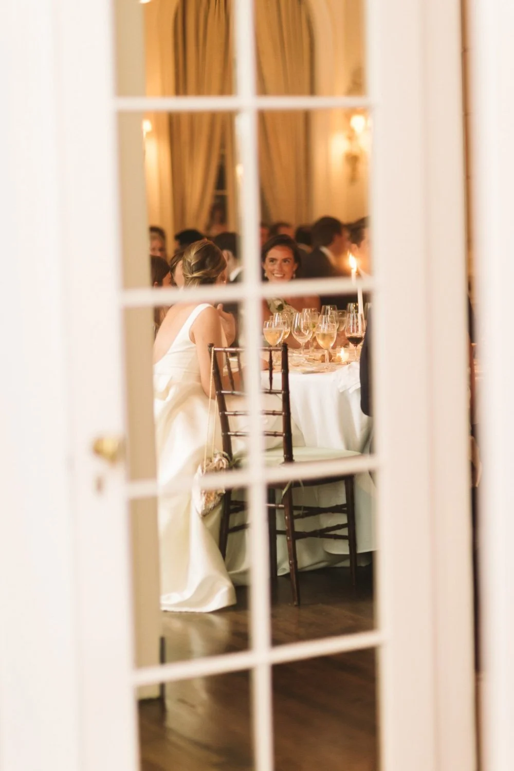 View through a window of a formal dinner at The Colony Club with women in dresses, glasses of wine, and candlelight.